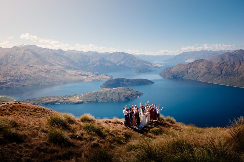 Wedding party gathered on a hillside overlooking a deep blue lake surrounded by mountains.