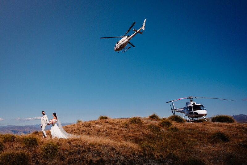 Bride and groom on a hillside with two helicopters nearby during an aerial wedding shoot.