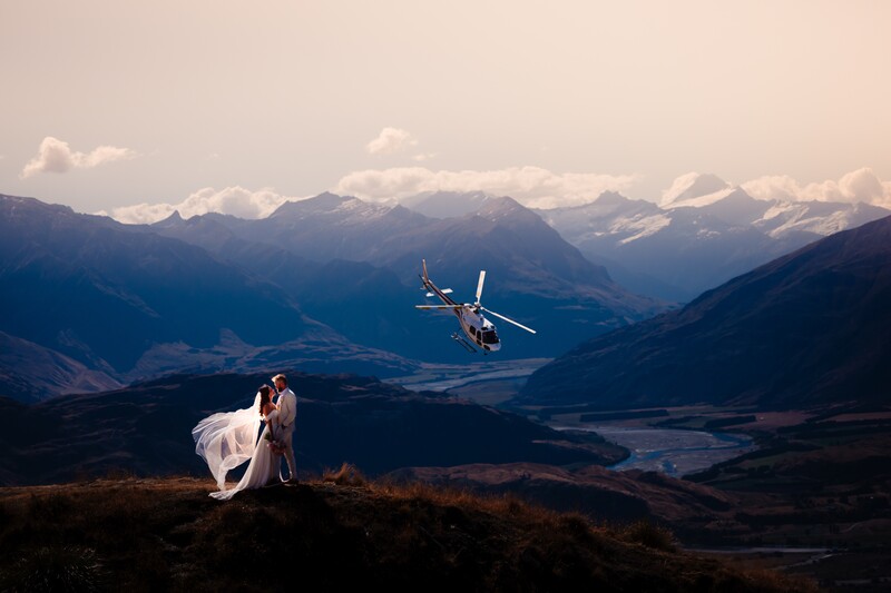 Bride in white dress standing on mountain ridge overlooking valley with helicopter in mid-air.