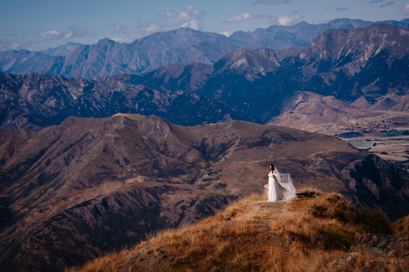 Bride in flowing white gown stands on arid hillside overlooking expansive mountain range under blue sky.