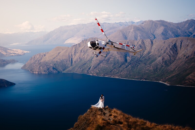 Helicopter descending toward a couple in wedding attire standing on a mountain ridge overlooking a deep blue alpine lake.