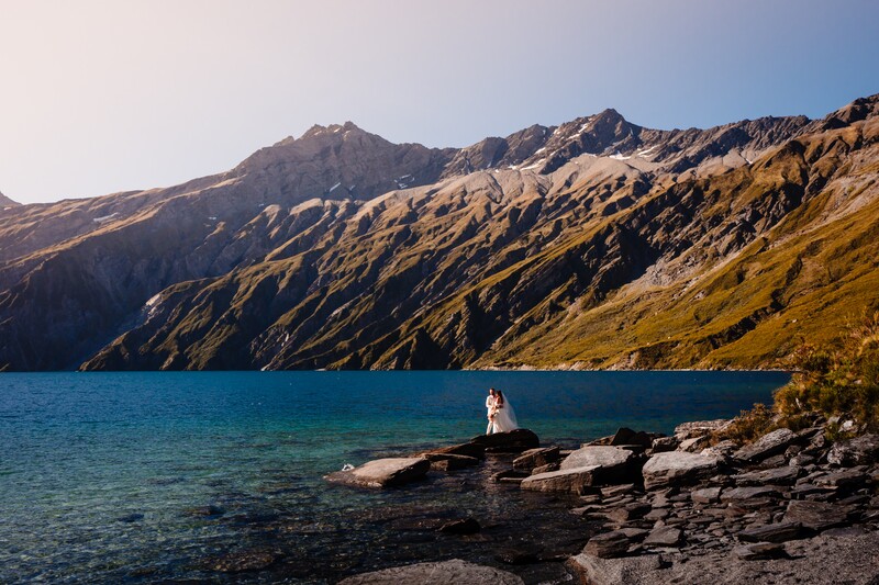 Person standing on rocks beside a turquoise alpine lake with steep brown mountains in background.