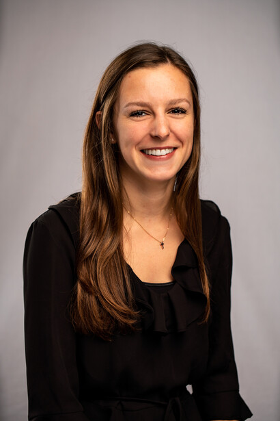 Woman with long dark hair smiling at camera wearing black blazer against neutral gray background.