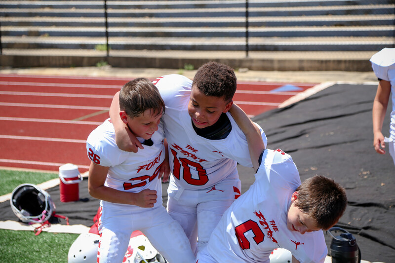 Three youth football players in white uniforms embrace on a stadium field during a game.