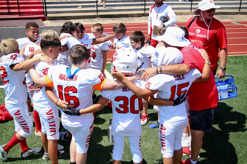 Youth football team huddles together on a field during practice or game with coaches nearby.