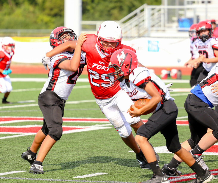 Youth football player in red jersey carrying ball while evading defenders during game.