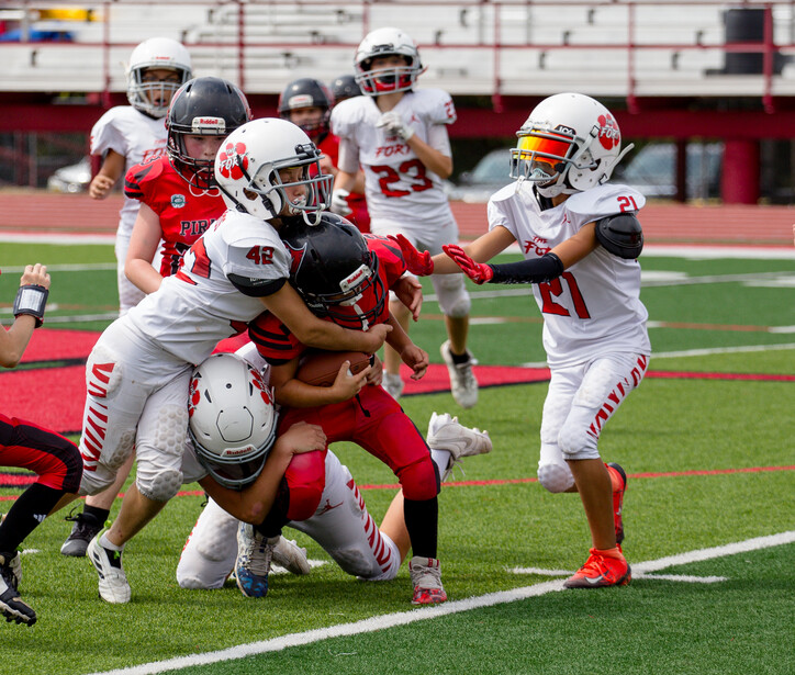 Youth football players in white and red uniforms engaged in a tackle during a game on an artificial turf field.