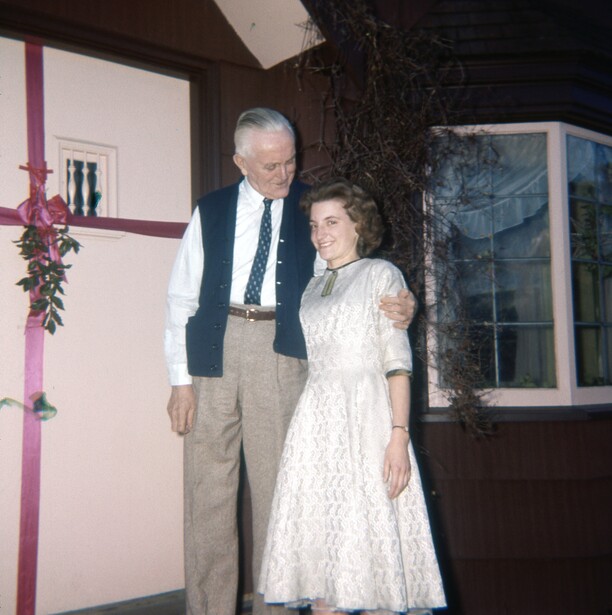 Older man in vest and tie standing beside woman in white lace dress indoors.