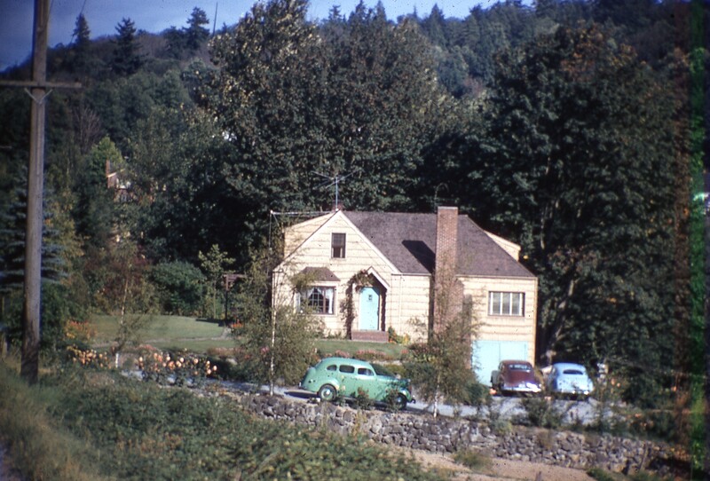 A cream-colored cottage with brick chimney nestled among evergreen trees, vintage car parked in front.