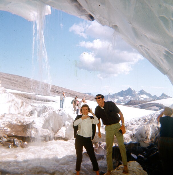 Group of hikers exploring beneath an ice overhang in a snowy alpine landscape with mountains visible in the distance.