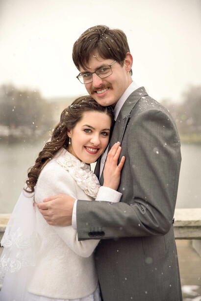 Couple posing together outdoors in falling snow near a body of water.