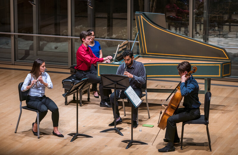 Four young musicians perform chamber music in a bright studio space with a grand piano illustration on the back wall.