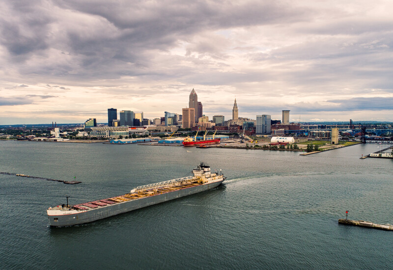 Large cargo ship anchored in harbor with city skyline and dramatic clouds in background.