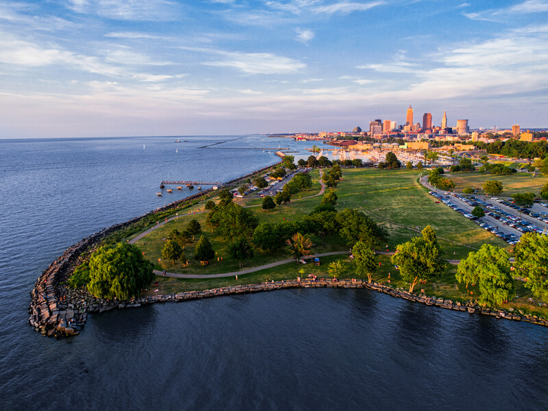 Aerial view of a waterfront peninsula with green parks and a city skyline across the bay at sunset.
