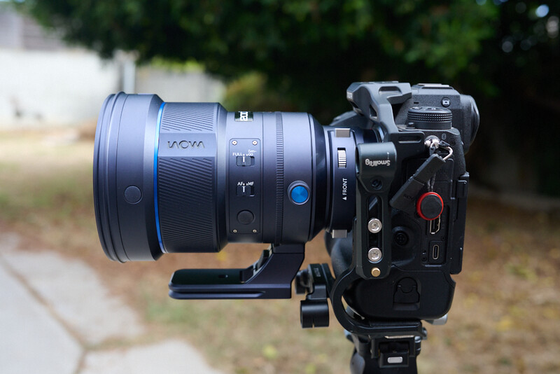 Cinema camera mounted on tripod with large telephoto lens attached outdoors.