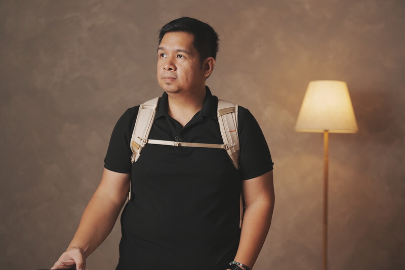 Man in black polo shirt with canvas backpack against warm studio backdrop.