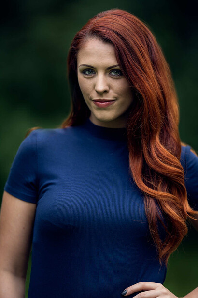 Woman with long reddish-brown hair wearing a dark blue short-sleeved shirt against a blurred green background.