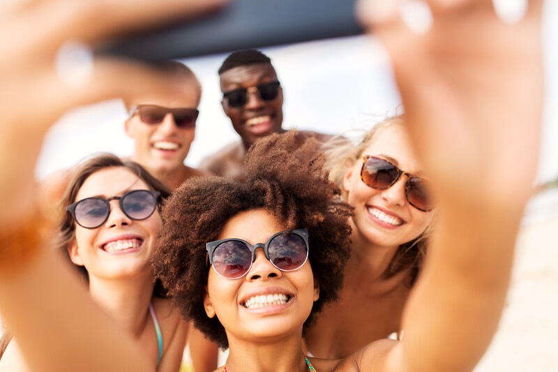 Group of five smiling people wearing sunglasses taking a selfie outdoors.