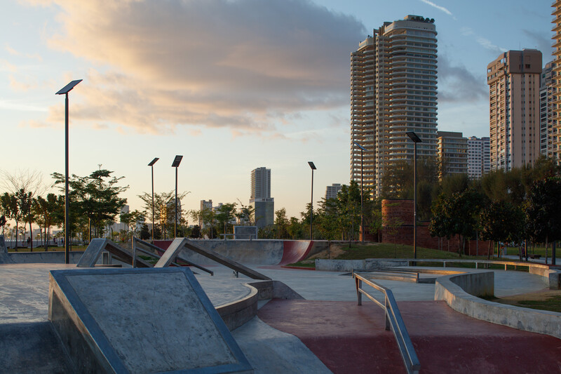 Skate park with concrete ramps and metal railings at golden hour, high-rise buildings in background.