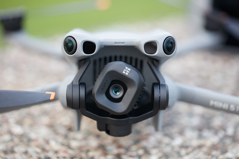 DJI Mini drone photographed from above, showing dual front cameras, sensors, and folded arms against gravel ground.