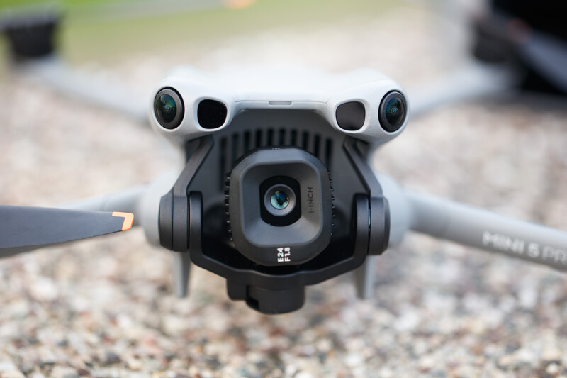DJI Mini drone hovering above gravel ground, showing front-facing camera, dual sensors, and articulated arms.