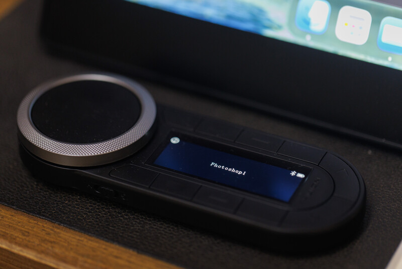 Close-up of a black wireless control pad with a large rotary knob and small display screen on a wooden surface.