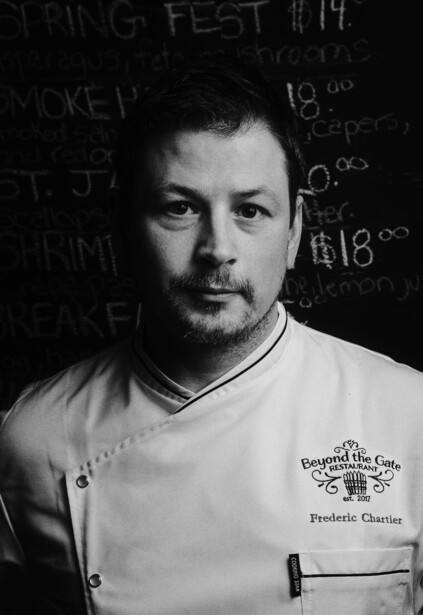 Black and white portrait of a man wearing a v-neck white t-shirt against a dark background with blurred text.