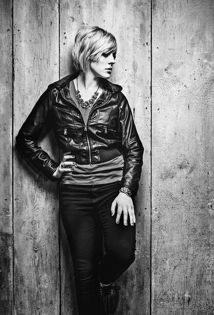 Person in black leather jacket and distressed pants against weathered wooden wall, looking to the side.