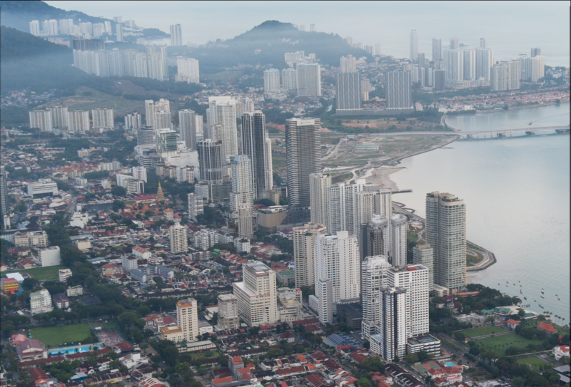 Aerial cityscape with dense high-rise towers along a waterfront, mountains in the distance, and lower-density residential areas in foreground.