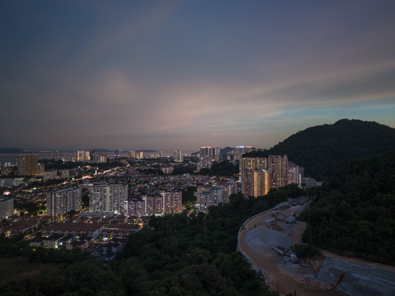 Urban landscape at dusk with illuminated high-rise apartments, a winding river reflecting twilight, and forested hillside.