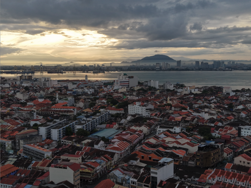 Aerial cityscape at dusk with red-roofed buildings, waterway, and distant mountains under cloudy sky.