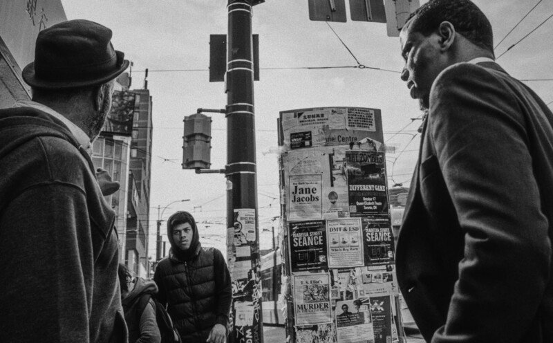 Three people on an urban street with posted advertisements on a pole behind them.