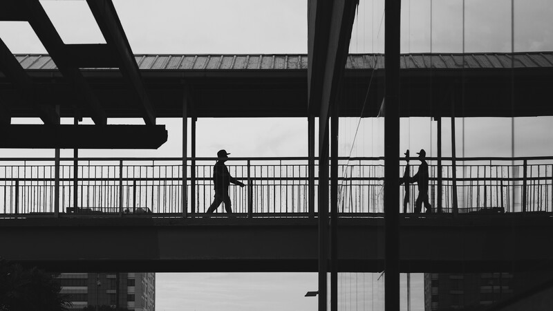 Silhouetted figures walking on an elevated pedestrian bridge with geometric structural elements and railings.