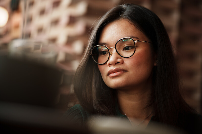 Woman wearing round-framed glasses gazes thoughtfully to the side against a brick wall backdrop.