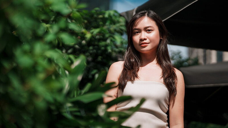 Young woman in light tank top posing among green foliage with natural sunlight.