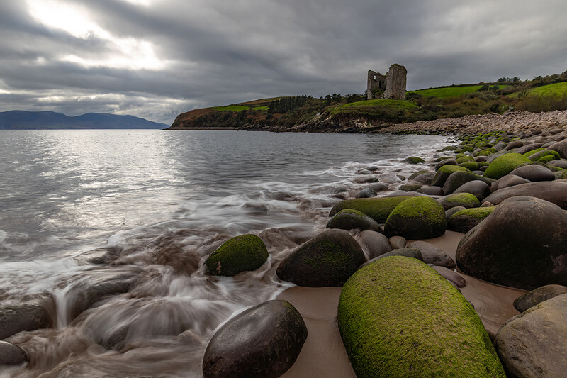 Moss-covered boulders on a rocky beach with waves and a ruined tower on a headland under dramatic clouds.