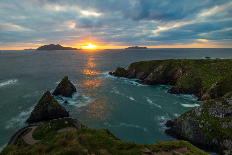 Dramatic coastal landscape with rocky outcrops and turquoise water at sunset, distant islands on horizon.
