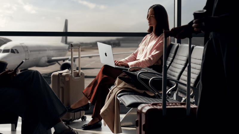 Woman working on laptop at airport window with airplane visible on tarmac.
