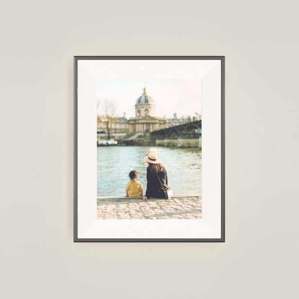 Two people sitting on a riverbank viewing a cityscape with a prominent clock tower across the water.