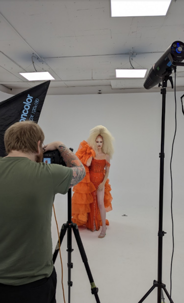 Photographer shooting a model in an orange dress in a studio with overhead lighting and a mounted camera on a crane or jib.