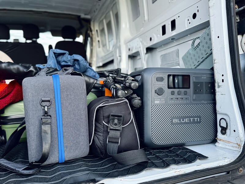 Interior of a van loaded with camera gear, including a gray camera bag with blue straps, black backpack, and a Bluetti power station.