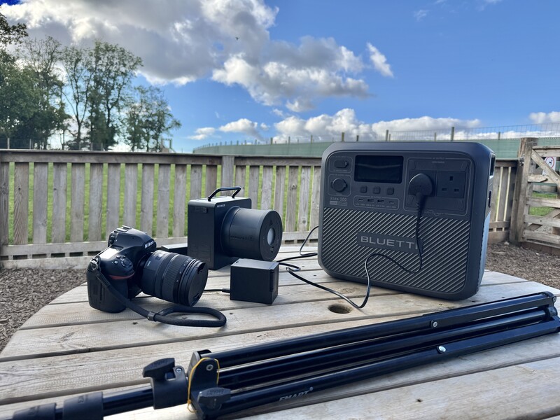 Photography equipment setup on wooden table outdoors, including camera, lens, portable speaker, and tripod rail.