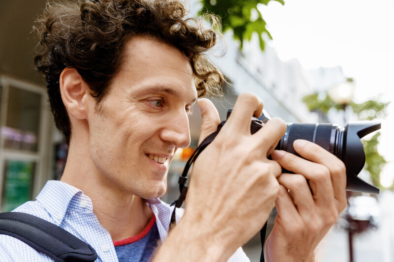 Male photographer holding a DSLR camera with telephoto lens, smiling while composing a shot outdoors.