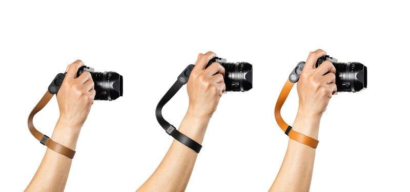 Three camera wrist straps in brown leather, black nylon, and orange nylon displayed on a hand holding a DSLR camera.
