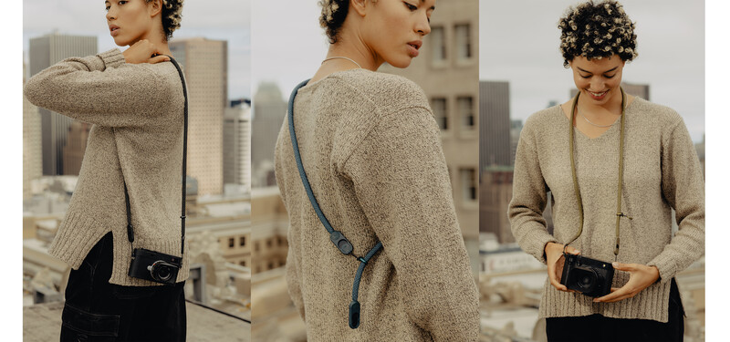 Woman in beige cardigan with black crossbody camera bag posed against urban skyline.