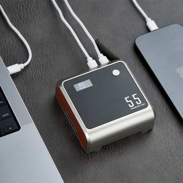 Compact portable power bank with digital display showing 55% battery level, connected via USB cables to laptops.
