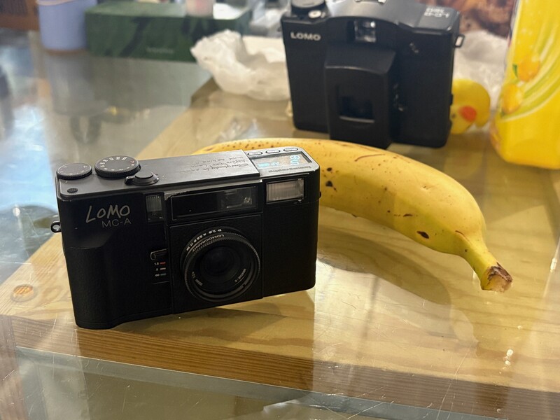 Black Lomo instant camera positioned next to a yellow banana on a wooden surface.