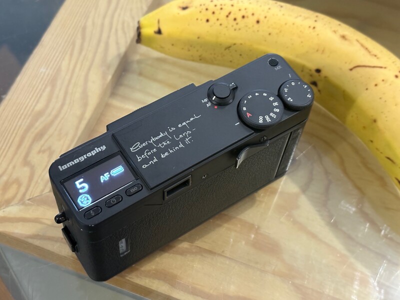 Compact digital camera with LCD screen and control dials positioned next to a yellow banana on wooden surface.