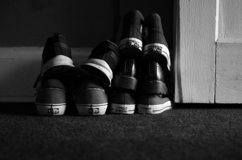 Four black sneakers with white stripe details arranged in a row against a dark floor and white wall.