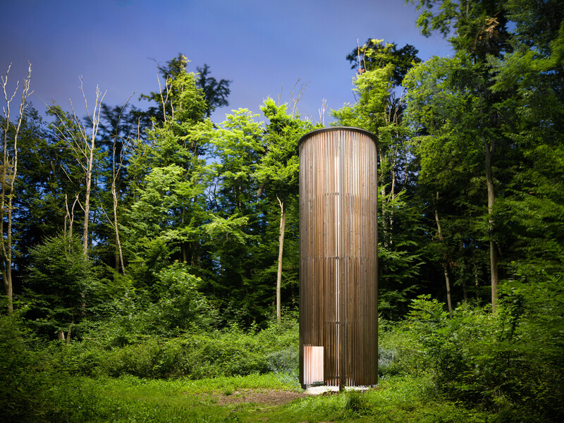 Tall cylindrical wooden structure with vertical paneling standing in a cleared grassy area surrounded by dense forest.